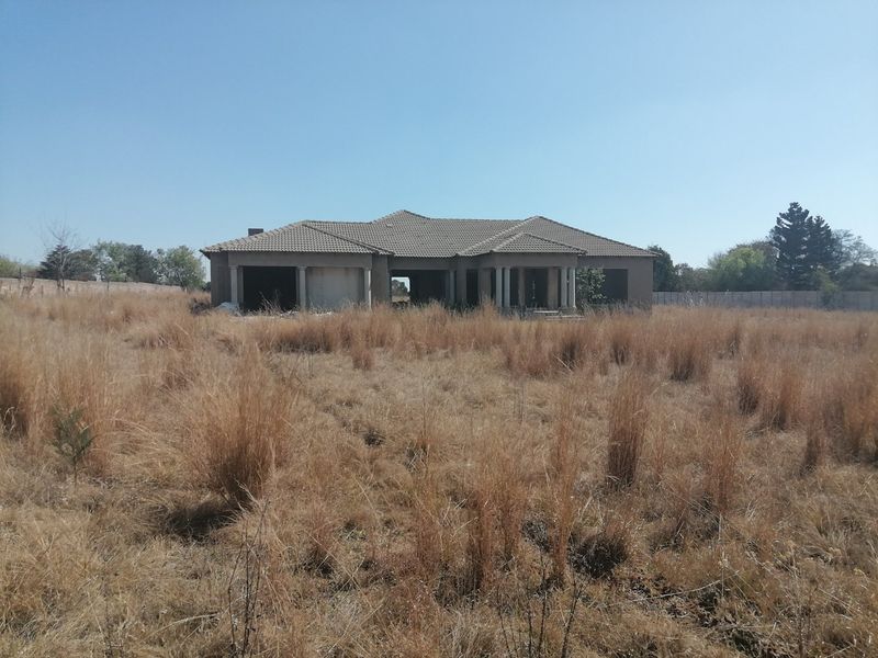 An unfinished residential property structure set on a large, undeveloped plot of land with dry grass and perimeter fencing.