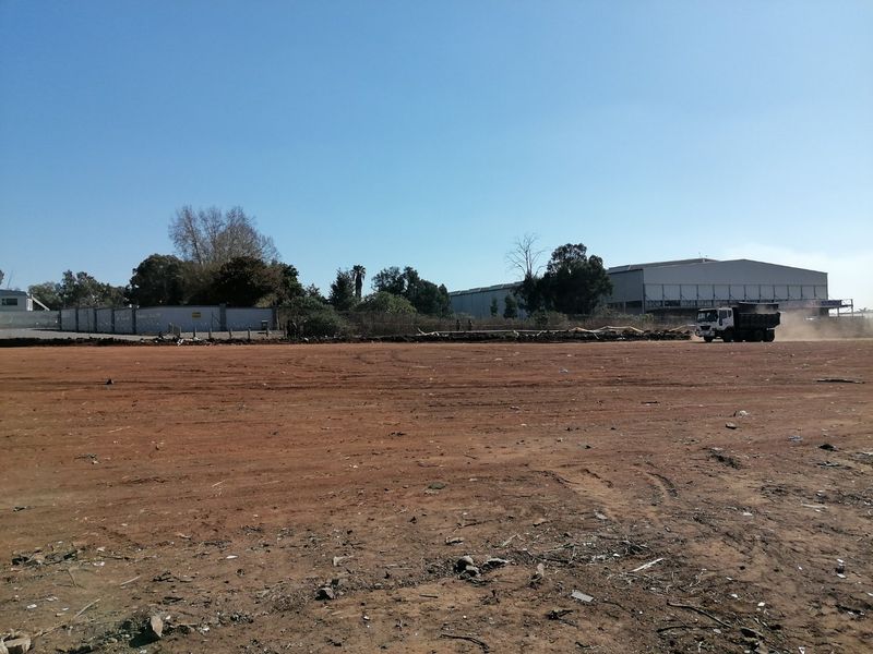 A wide-angle view of a large, cleared residential vacant land plot with perimeter walling and surrounding industrial infrastructure.
