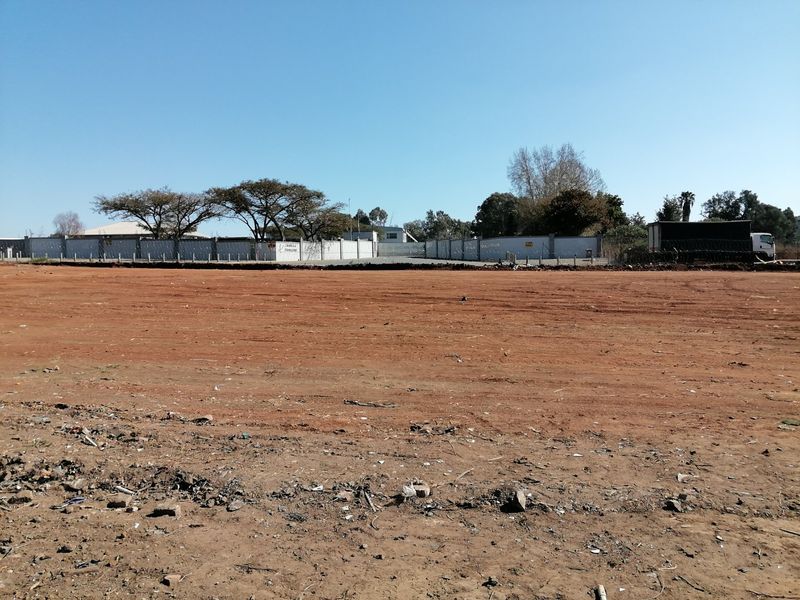 A wide view of a vacant residential plot featuring cleared red earth and a perimeter wall with security fencing.