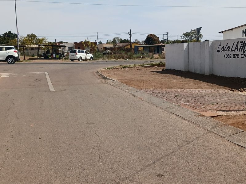 Street view showing the property boundary wall, paved driveway entrance, and adjacent asphalt road infrastructure.
