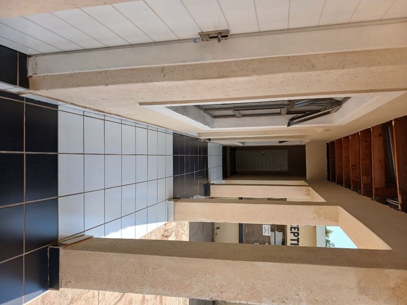 A covered outdoor walkway or patio area featuring tiled flooring, concrete support pillars, and exposed timber rafters, requiring maintenance and finishing.
