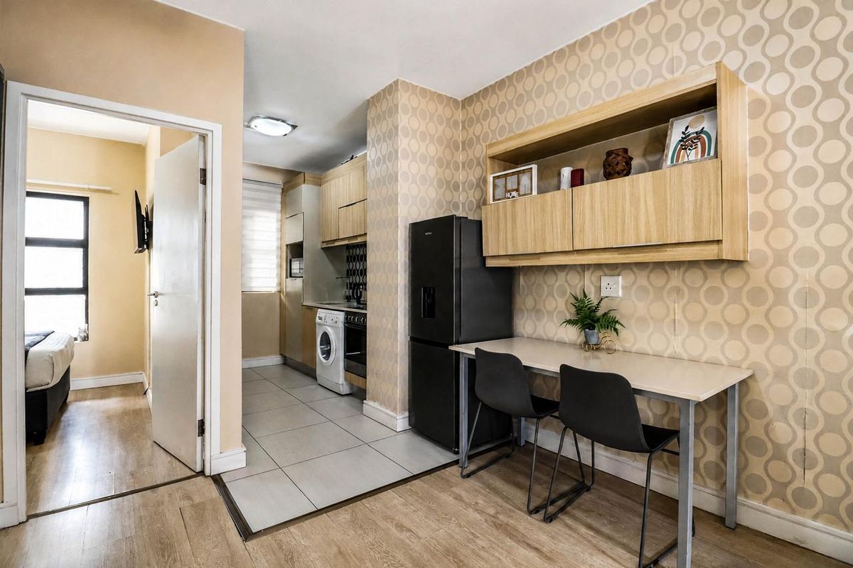 Open-plan kitchen and dining area featuring tiled and laminate flooring, built-in cabinetry, and integrated kitchen appliances, with an adjacent bedroom visible.