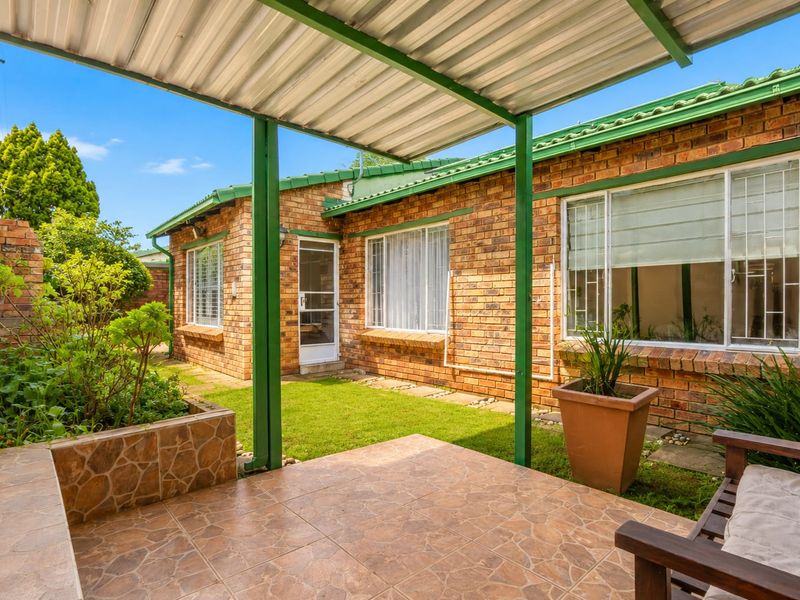 A covered patio area featuring tiled flooring, a corrugated metal roof, and a raised stone planter box, overlooking a small garden space.