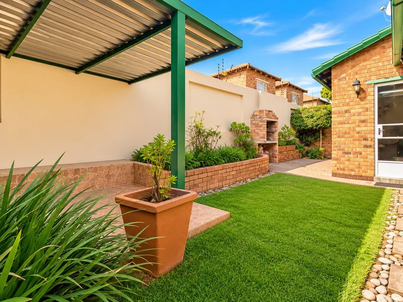 A well-maintained rear garden featuring a covered patio, built-in braai, brick boundary walls, and a neat lawn area.