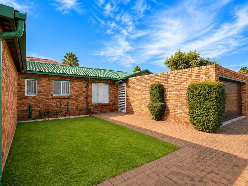 A well-maintained townhouse exterior featuring a face brick facade, green tiled roof, paved driveway, and a private front garden area.