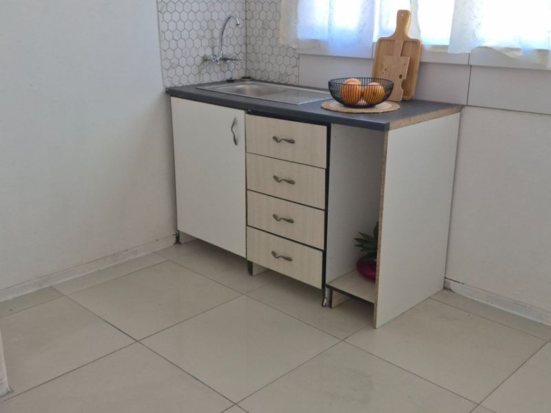 A compact kitchen area featuring a small sink unit, tiled backsplash, and basic cabinetry with an open storage section.