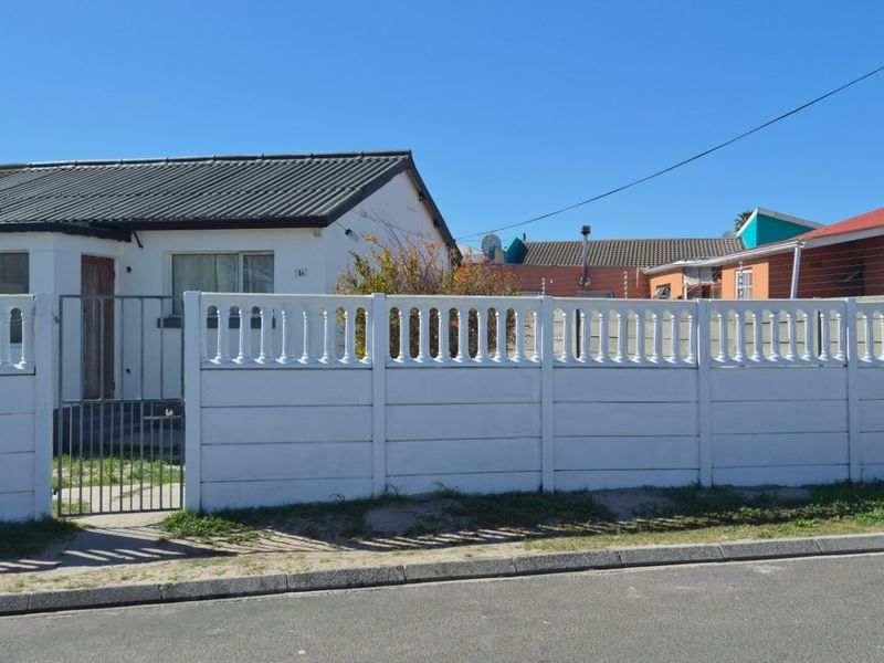 Front view of a residential property featuring a white precast concrete boundary wall, metal gate, and corrugated metal roof with perimeter electric fencing.