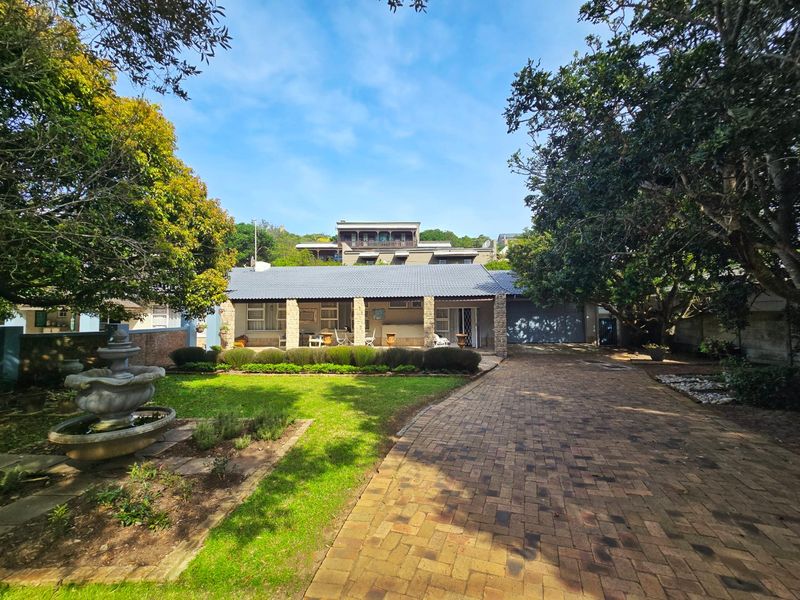 Front view of the property featuring a brick paved driveway, stone-pillared covered patio, landscaped garden with a fountain, and a garage.