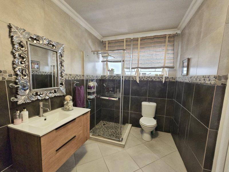 A well-maintained bathroom featuring a modern wood-finish vanity, a glass-enclosed shower, and neutral floor-to-ceiling tiling with a decorative pebble accent border.