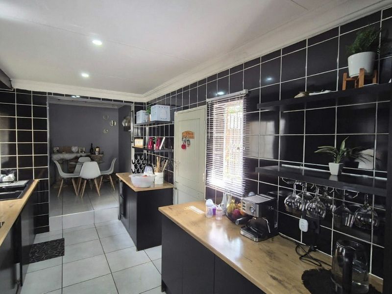 Modern kitchen featuring black tiled walls, wooden countertops, built-in appliances, and open shelving, with an adjacent dining area visible.