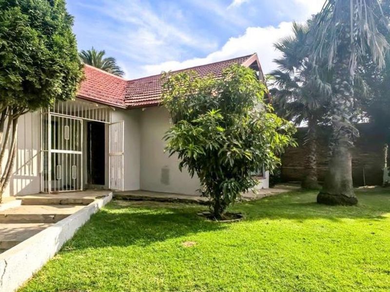 A view of the rear garden featuring a lawn, mature trees, and a paved entrance with a security gate.