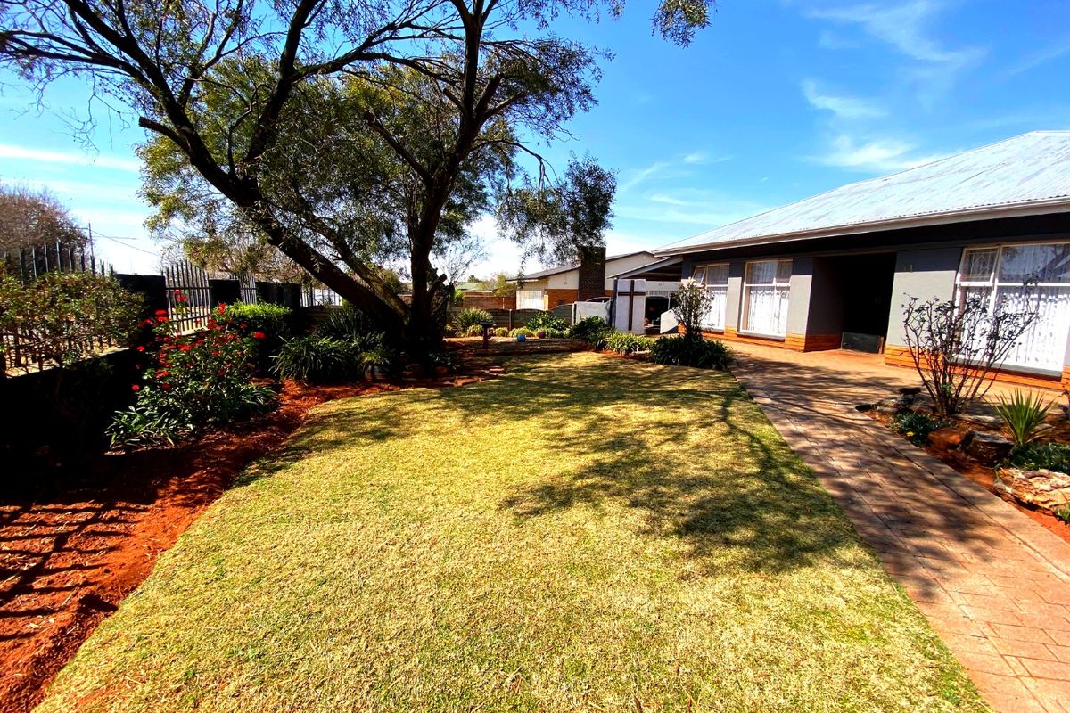 A well-maintained rear garden featuring a paved walkway, established trees, and a clear view of the house exterior with a metal roof.