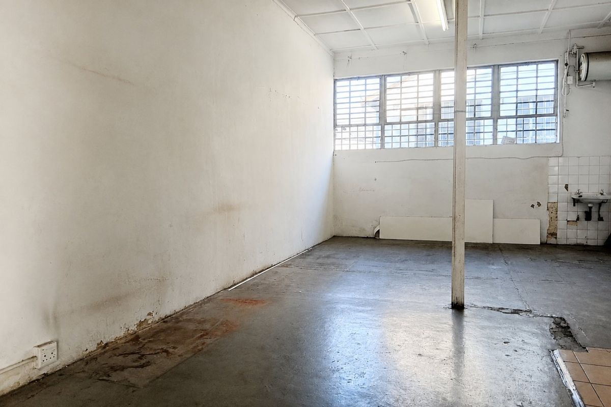 An empty commercial utility space featuring concrete flooring, a wall-mounted basin, and large windows with security bars, requiring cosmetic refurbishment.