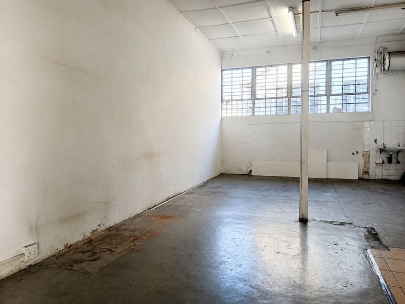 An empty commercial utility space featuring concrete flooring, a wall-mounted basin, and large windows with security bars, requiring cosmetic refurbishment.