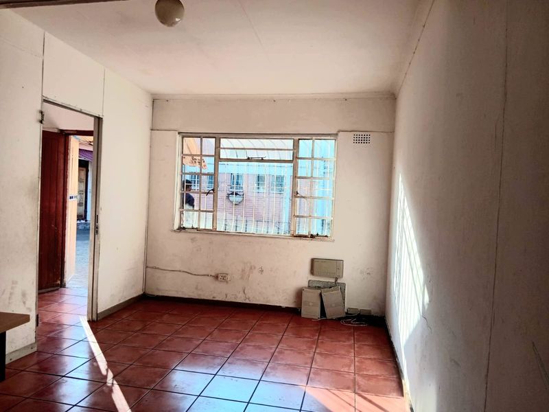 An empty commercial office space featuring tiled flooring, a steel-framed window with security bars, and plain white walls in need of refurbishment.