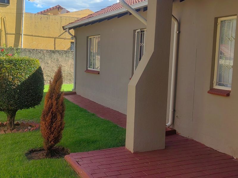 A rear exterior view of the property featuring a paved brick patio, sliding glass doors with security gates, and a landscaped garden area.
