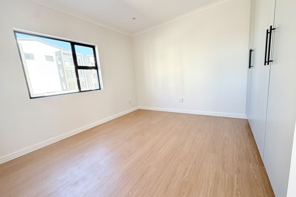 A bright, empty bedroom featuring modern laminate flooring, built-in cupboards, and a large aluminium-framed window providing natural light.