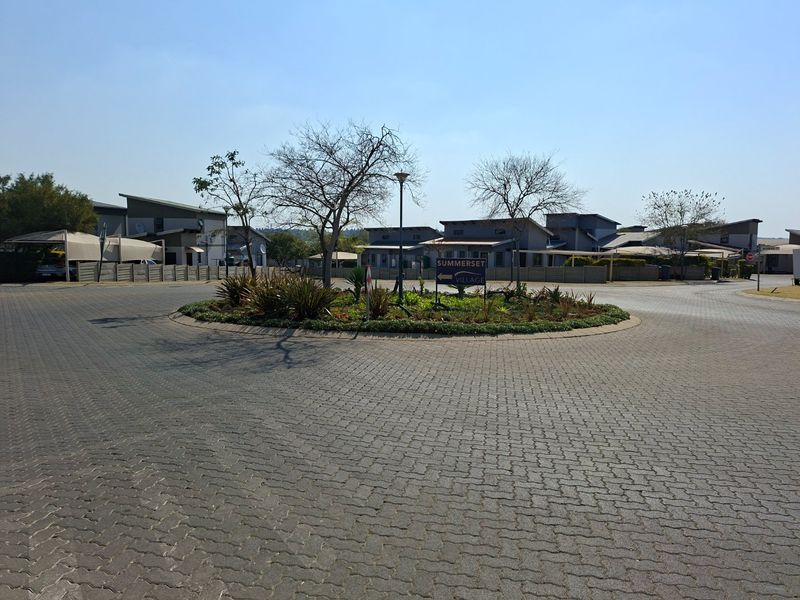 A view of the paved roundabout and entrance area of Summerset Village, featuring landscaped central island and established residential buildings in the background.