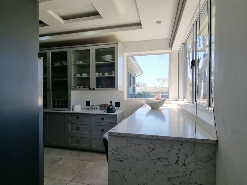 Modern kitchen featuring elegant grey cabinetry, stone countertops, and a bright, airy atmosphere with large windows and recessed ceiling details.