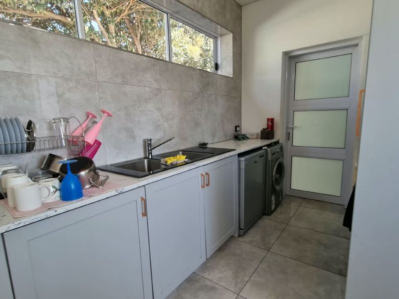 A functional scullery featuring modern grey cabinetry, stone-look countertops, a stainless steel sink, and tiled finishes with convenient access to laundry appliances.
