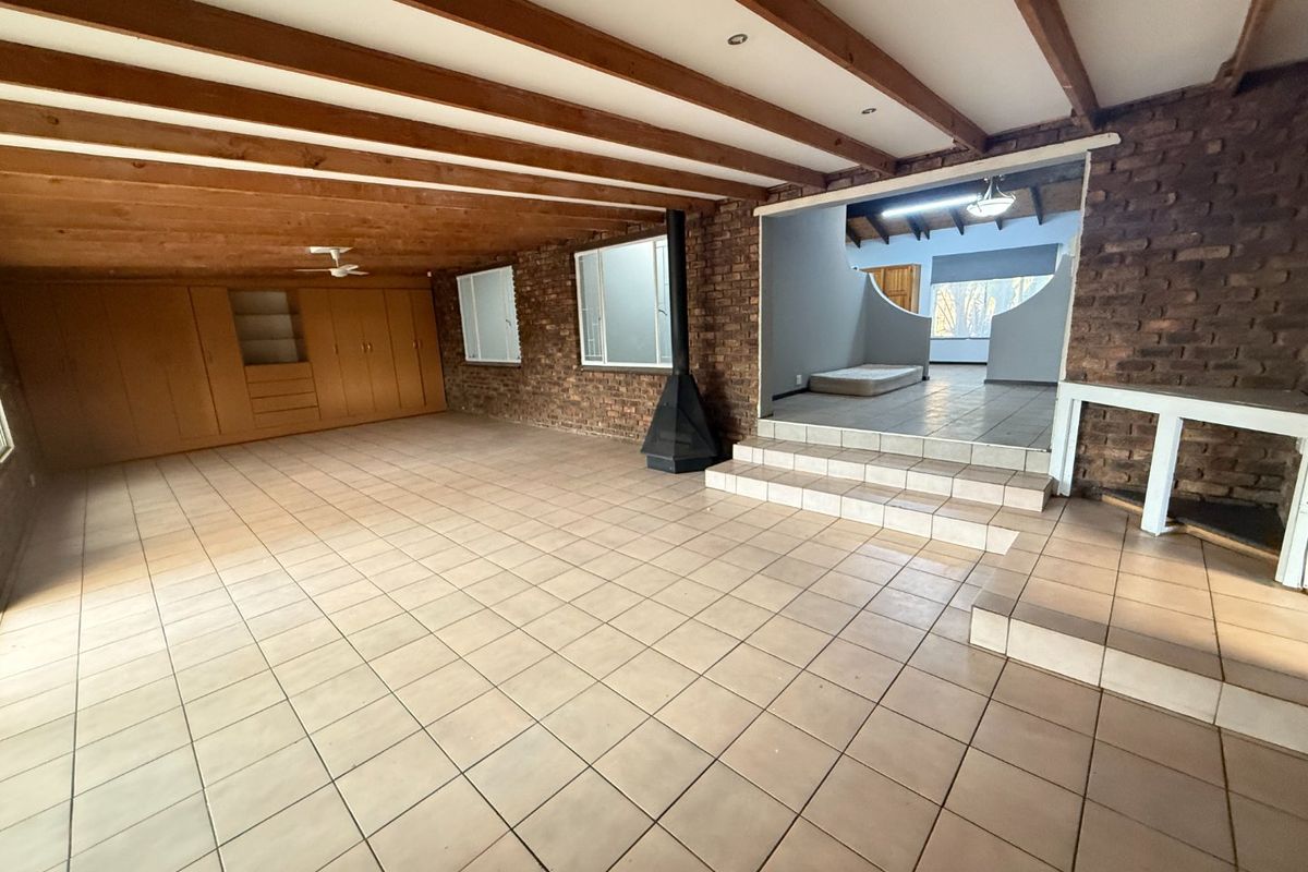 Spacious living area featuring exposed timber beams, brick walls, tiled flooring, and a freestanding fireplace, leading to an elevated adjacent room.