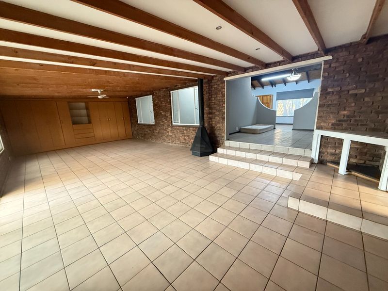 Spacious living area featuring exposed timber beams, brick walls, tiled flooring, and a freestanding fireplace, leading to an elevated adjacent room.