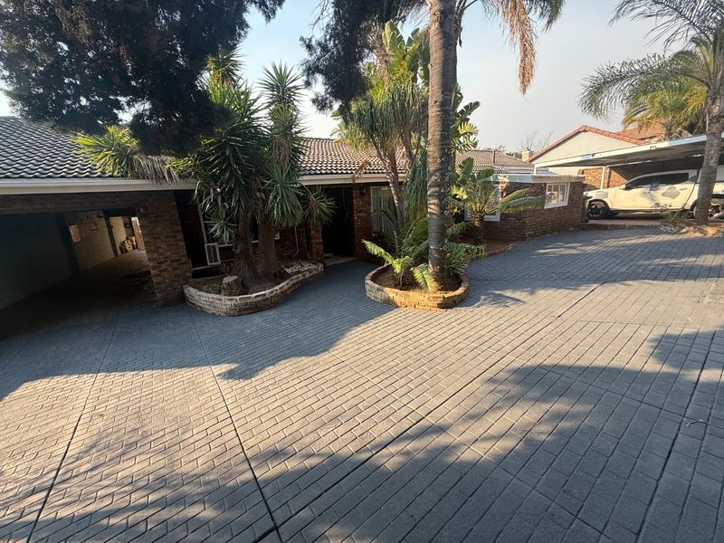 Front exterior view of the house featuring a paved driveway, brick facade, tiled roof, and established garden beds with a carport.