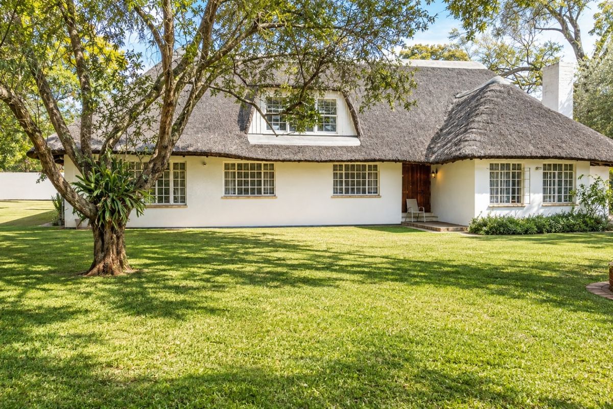 A charming house exterior featuring a traditional thatched roof, white facade, and grid-pattern windows, set within a spacious, well-maintained garden.