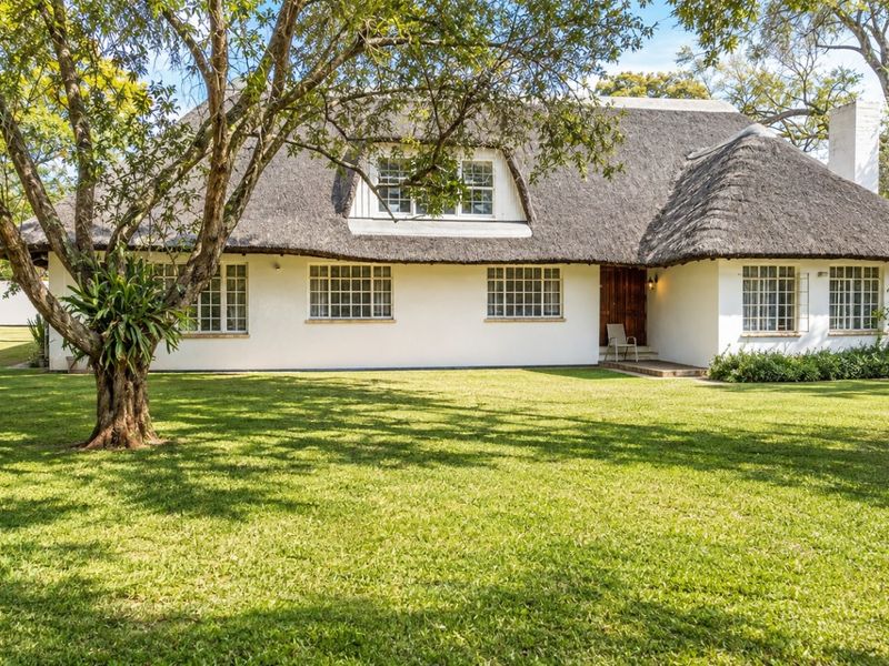 A charming house exterior featuring a traditional thatched roof, white facade, and grid-pattern windows, set within a spacious, well-maintained garden.
