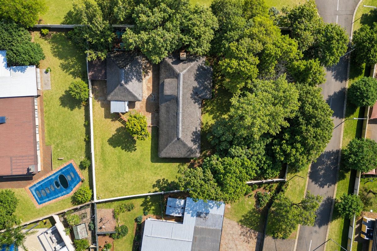 Aerial view of a residential property featuring a main house with a thatched roof, a swimming pool, and mature trees within a fenced garden.