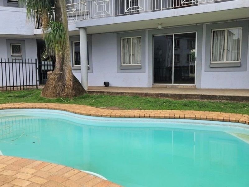 Exterior view of an apartment complex featuring a swimming pool with brick coping, a concrete patio area, and sliding glass door access.