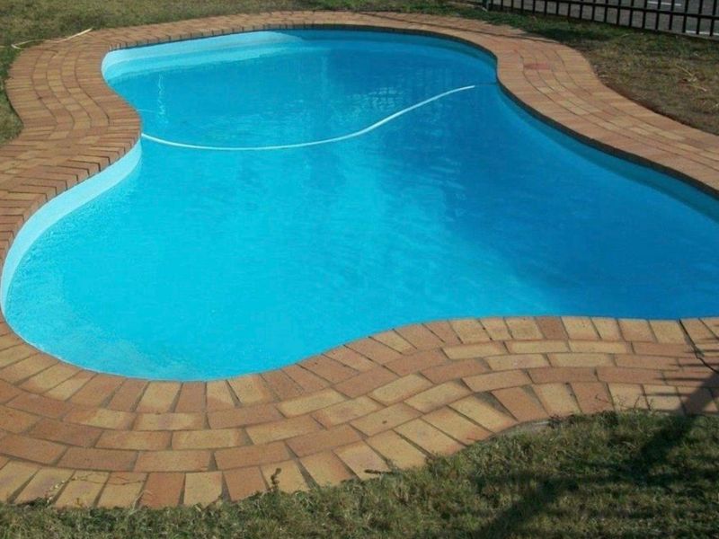 A kidney-shaped swimming pool featuring a clean brick-paved surround, set within a residential garden area.
