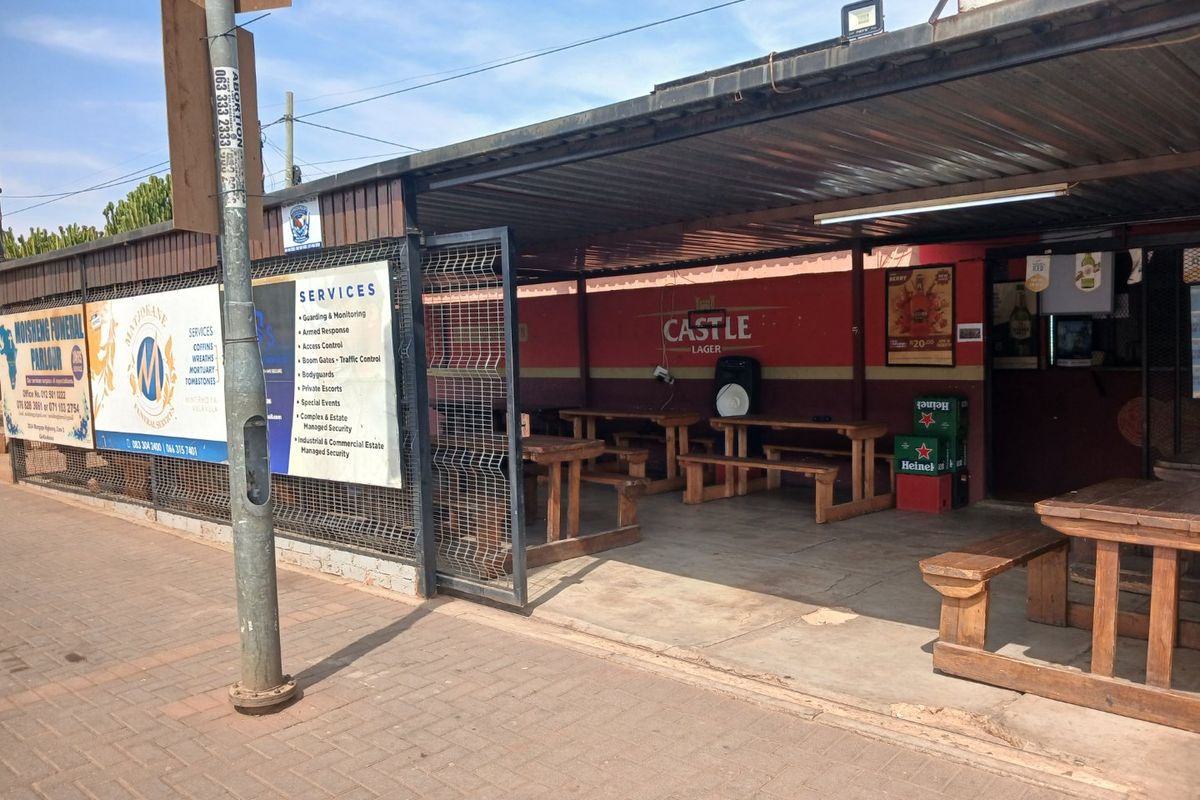 A covered outdoor patio area featuring a corrugated metal roof, concrete flooring, and a metal security gate, currently utilized as a commercial seating space.