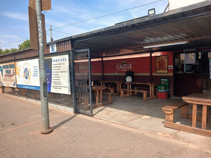 A covered outdoor patio area featuring a corrugated metal roof, concrete flooring, and a metal security gate, currently utilized as a commercial seating space.