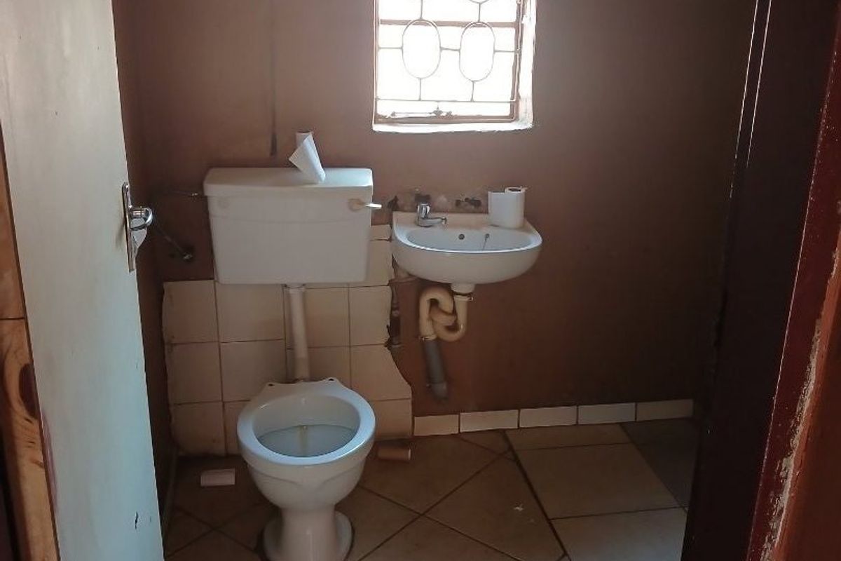 A compact bathroom featuring a ceramic toilet, wall-mounted basin, and tiled flooring, currently requiring cosmetic updates and repairs to wall finishes and plumbing.