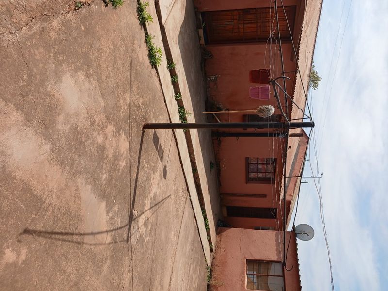 Rear exterior view of the property featuring concrete paving, weathered exterior walls, and metal window frames, showing significant signs of wear and aging.