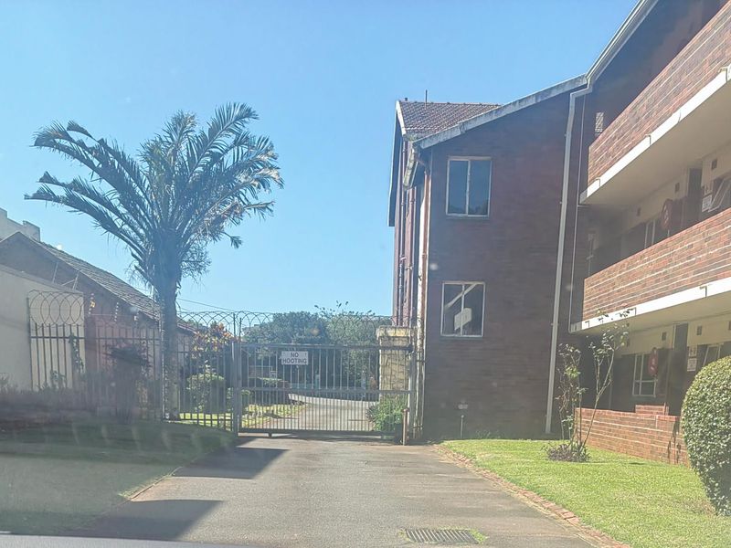 Exterior view of an apartment complex entrance featuring a brick facade, security gate, and paved driveway access.