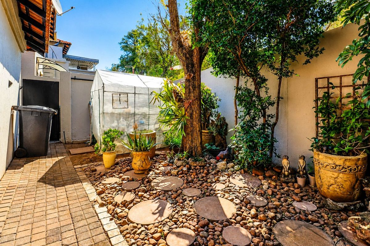A private rear courtyard area featuring a paved walkway, landscaped garden bed with stone mulch, and established trees against a high boundary wall.