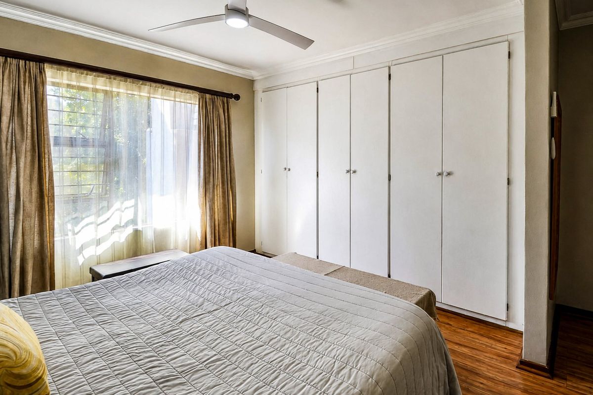 A well-maintained bedroom featuring laminate flooring, a ceiling fan, and extensive built-in cupboard storage with a large window allowing natural light.