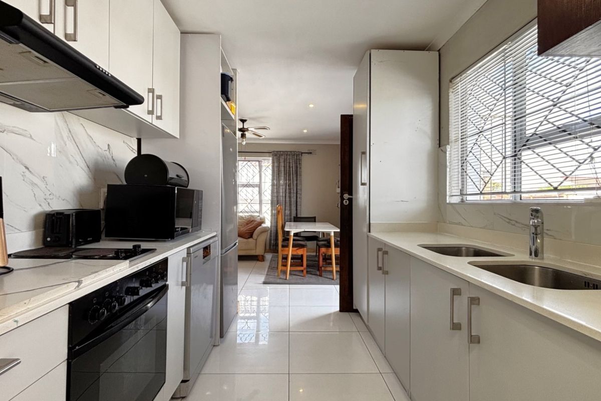 Modern galley-style kitchen featuring white cabinetry, stone countertops, integrated oven, extractor fan, and double basin sink with polished tiled flooring.