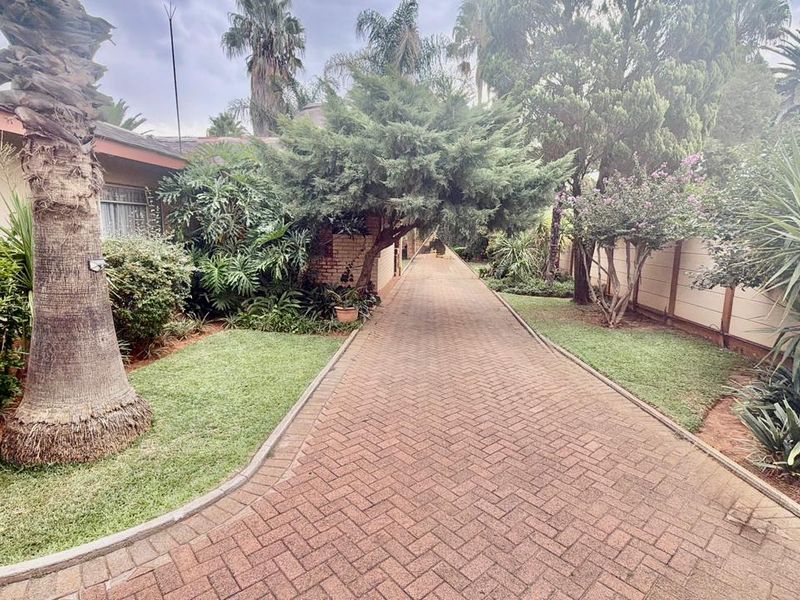A well-maintained paved driveway leading towards the property, flanked by established garden beds and mature trees.