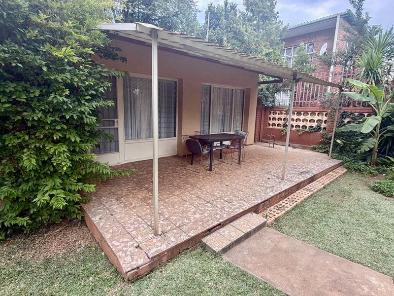 A covered outdoor patio area featuring tiled flooring and sliding glass doors, overlooking a private garden space with established landscaping.