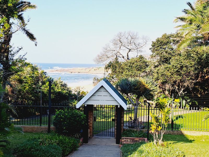 A scenic view of the coastline featuring a gated entrance with a pitched roof structure, surrounded by landscaped gardens and perimeter fencing.