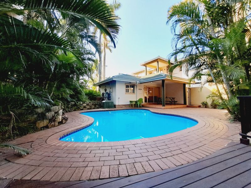 A well-maintained outdoor pool area featuring a curved swimming pool, brick paved surround, and a covered patio area set within a landscaped garden.