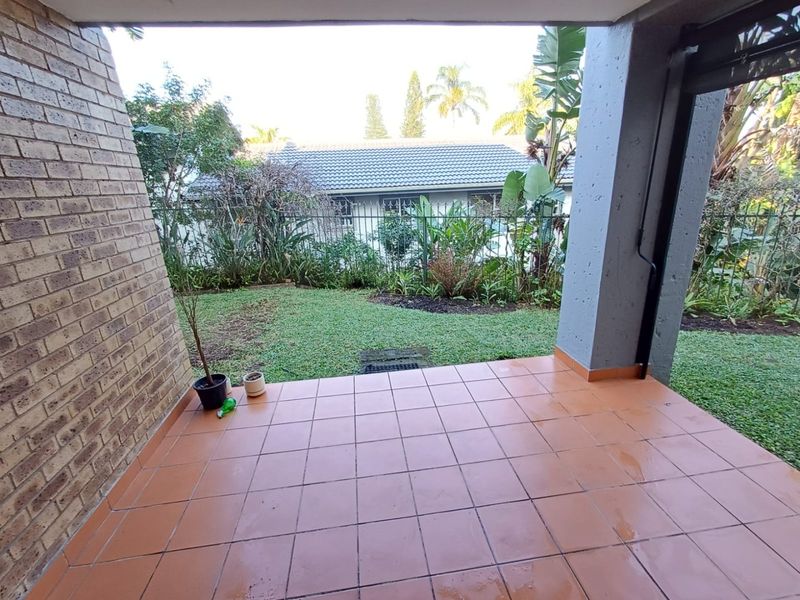 A covered patio area with tiled flooring, overlooking a private garden space with established greenery and a perimeter security fence.