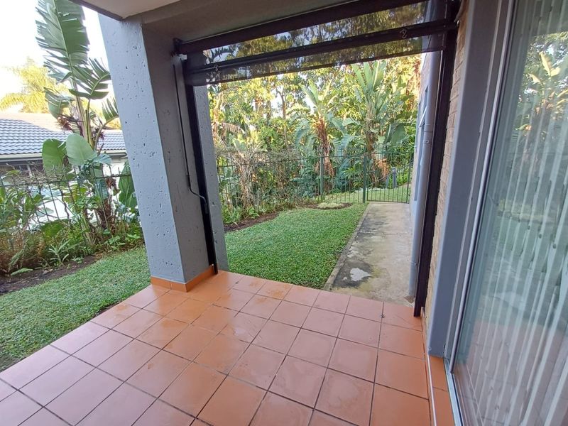 A covered patio area featuring tiled flooring, a retractable blind, and direct access to a private garden space through sliding glass doors.