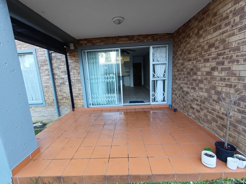 A covered patio area featuring tiled flooring, brick walls, and a sliding glass door with security gate access to the interior.
