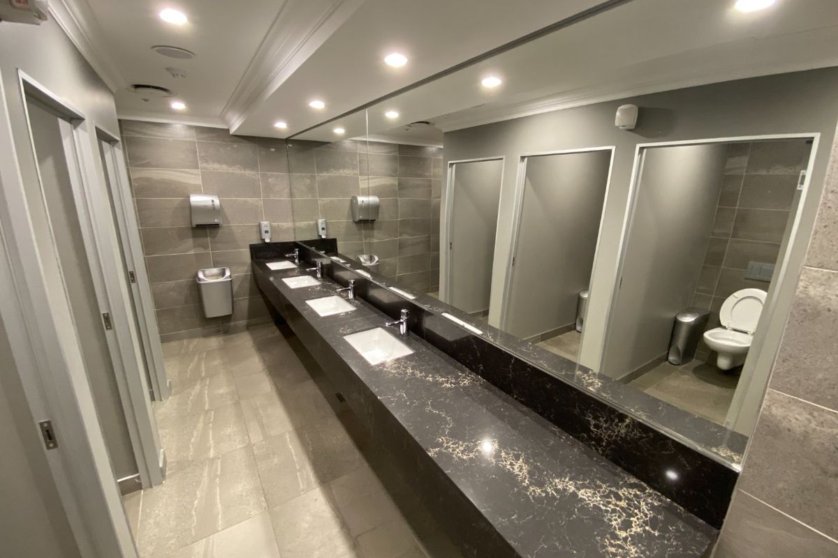 Modern commercial bathroom featuring a long stone vanity with multiple undermount sinks, large wall-to-wall mirror, and private toilet cubicles with tiled finishes.