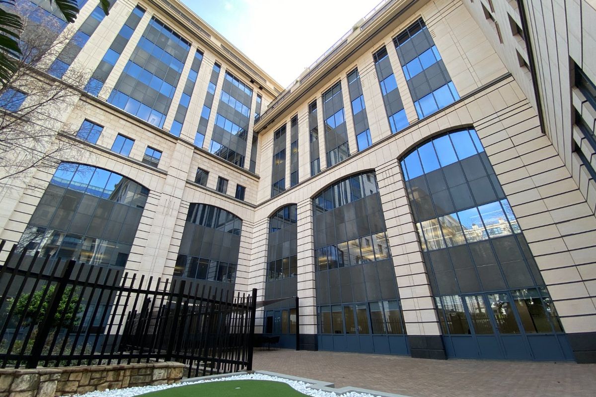 A modern commercial building exterior featuring a stone facade, large glass windows, and a paved courtyard area with security fencing.