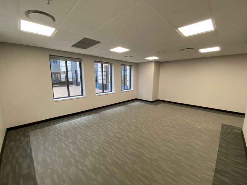 A bright, vacant commercial office space featuring neutral carpet tiles, a suspended ceiling with recessed lighting, and large windows providing natural light.